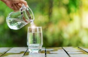 Pouring clean water from a jug into a glass placed on a wooden bar.